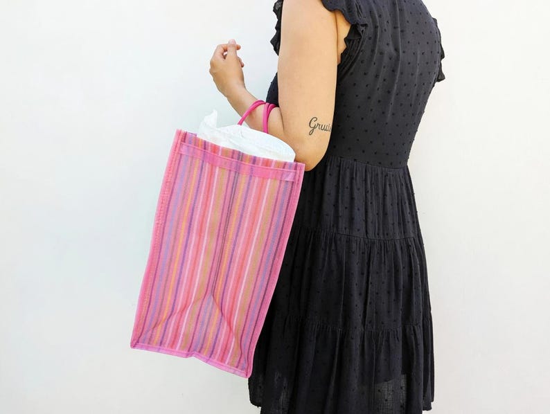 May include: A rectangular tote bag featuring vertical stripes in shades of pink, purple, orange, and white. The bag has two pink circular handles and is constructed from a semi-transparent material, revealing a white interior lining inside.