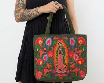 Può includere: Una borsa tote verde oliva con vivaci ricami floreali multicolori che circondano una figura centrale della Vergine di Guadalupe. Il design presenta fiori rosa brillante, arancioni e gialli con foglie verdi su una base di tessuto scuro.
