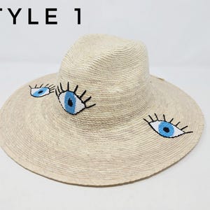 May include: A straw hat with a wide brim and three embroidered blue eyes. The hat is light beige and has a dark brown band around the crown.