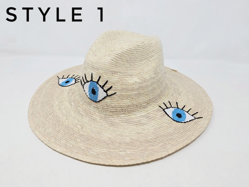 May include: A straw hat with a wide brim and three embroidered blue eyes. The hat is light beige and has a dark brown band around the crown.