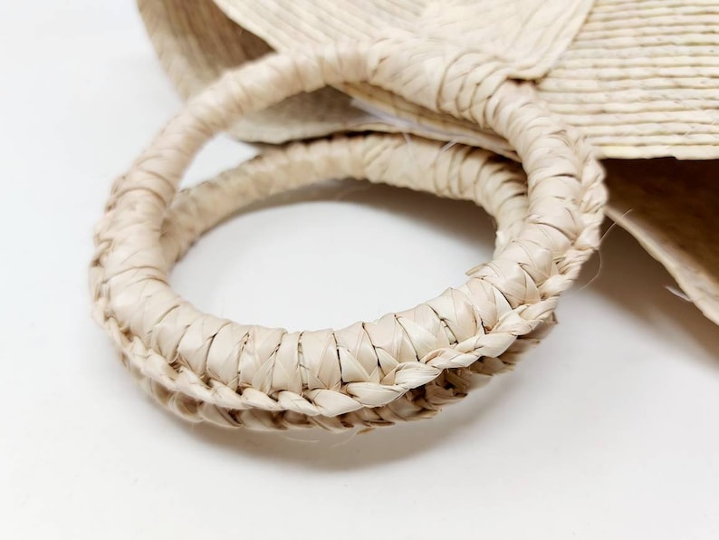 May include: A circular handle made of woven light beige straw material. The handle features a tightly braided texture and is attached to a larger woven straw bag body. The natural fibers show a consistent pale tan color throughout the structure.