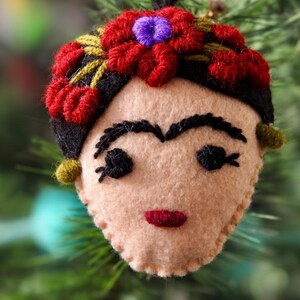 May include: A felt ornament shaped like a face with black embroidered eyebrows, eyes, and a red mouth. The figure has black hair adorned with red and purple embroidered flowers and small green circular accents on the sides of the head.