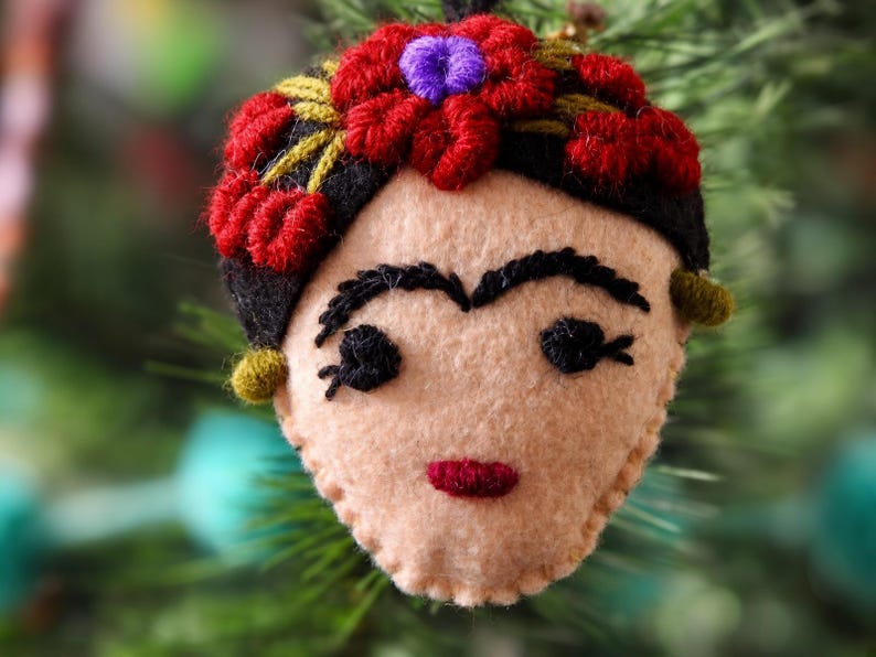 May include: A felt ornament shaped like a face with black embroidered eyebrows, eyes, and a red mouth. The figure has black hair adorned with red and purple embroidered flowers and small green circular accents on the sides of the head.