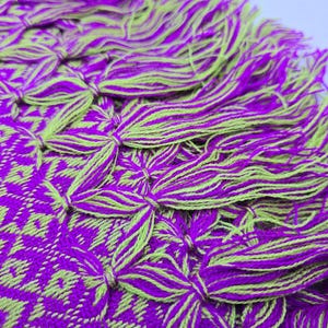 May include: A woven textile featuring a vibrant purple and lime green geometric diamond pattern. The edge of the fabric is finished with a series of thick, knotted tassels that alternate between the same purple and lime green yarn colors.