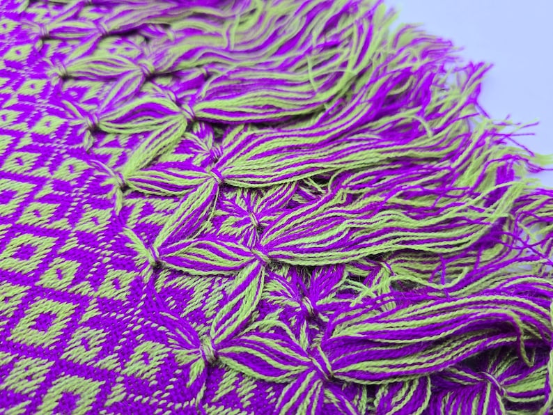 May include: A woven textile featuring a vibrant purple and lime green geometric diamond pattern. The edge of the fabric is finished with a series of thick, knotted tassels that alternate between the same purple and lime green yarn colors.