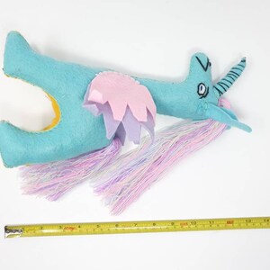 Puede incluir: Un peluche de unicornio de fieltro turquesa con la parte inferior amarilla, alas superpuestas rosas y moradas, y una larga melena y cola de hilo multicolor. Una cinta m&eacute;trica amarilla debajo muestra una longitud de unos 40,6 cm.