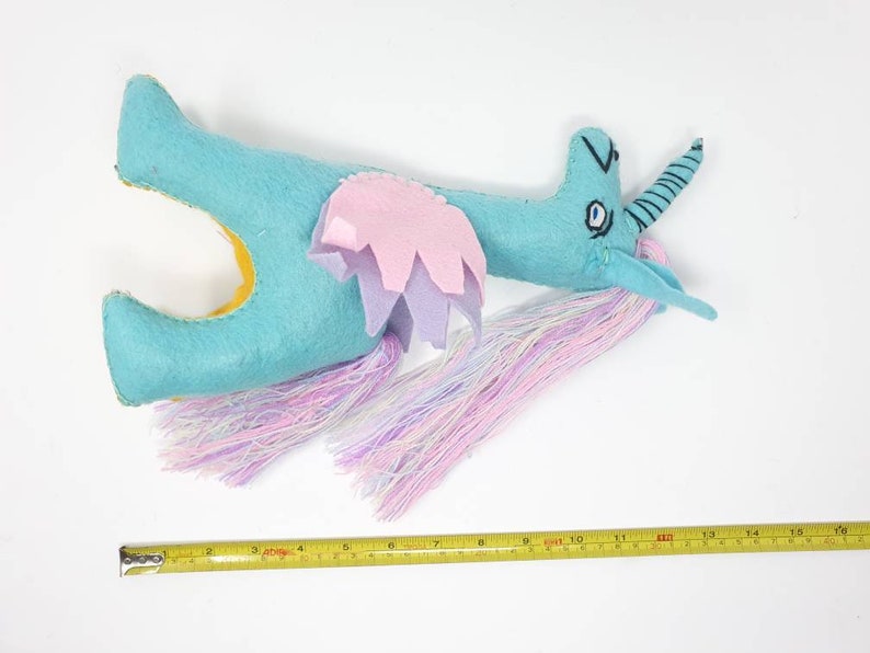 Puede incluir: Un peluche de unicornio de fieltro turquesa con la parte inferior amarilla, alas superpuestas rosas y moradas, y una larga melena y cola de hilo multicolor. Una cinta m&eacute;trica amarilla debajo muestra una longitud de unos 40,6 cm.