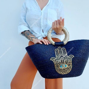 以下が含まれることがあります： 黒い籐製のトートバッグで、大きな金のハンズのデザインと青い目があります。バッグには自然な籐製のハンドルが付いています。