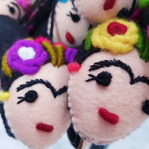 May include: A collection of handcrafted felt ornaments shaped like faces with black hair, embroidered eyebrows, red lips, and colorful floral accents in shades of purple, yellow, and green. Each piece features stitched details and a soft texture.