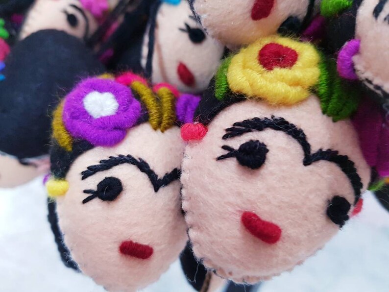 May include: A collection of handcrafted felt ornaments shaped like faces with black hair, embroidered eyebrows, red lips, and colorful floral accents in shades of purple, yellow, and green. Each piece features stitched details and a soft texture.