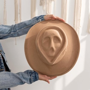 May include: A tan felt hat featuring a moulded crown shaped like a human face with recessed eye sockets and a mouth opening. The hat has a wide, flat brim and a smooth, uniform texture throughout the entire surface of the tan material.