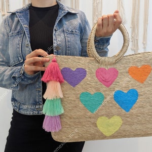 May include: A rectangular woven straw tote bag featuring two round top handles and a multi-colored tassel charm. The front of the bag is decorated with six embroidered heart shapes in purple, pink, orange, teal, blue, and yellow.