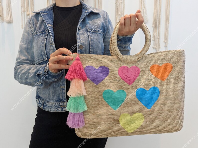 May include: A rectangular woven straw tote bag featuring two round top handles and a multi-colored tassel charm. The front of the bag is decorated with six embroidered heart shapes in purple, pink, orange, teal, blue, and yellow.