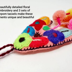 May include: Pink felt letter A with colourful floral embroidery and three sets of pompom tassels. The letter is decorated with bright pink, red, blue, green, yellow, and white flowers.
