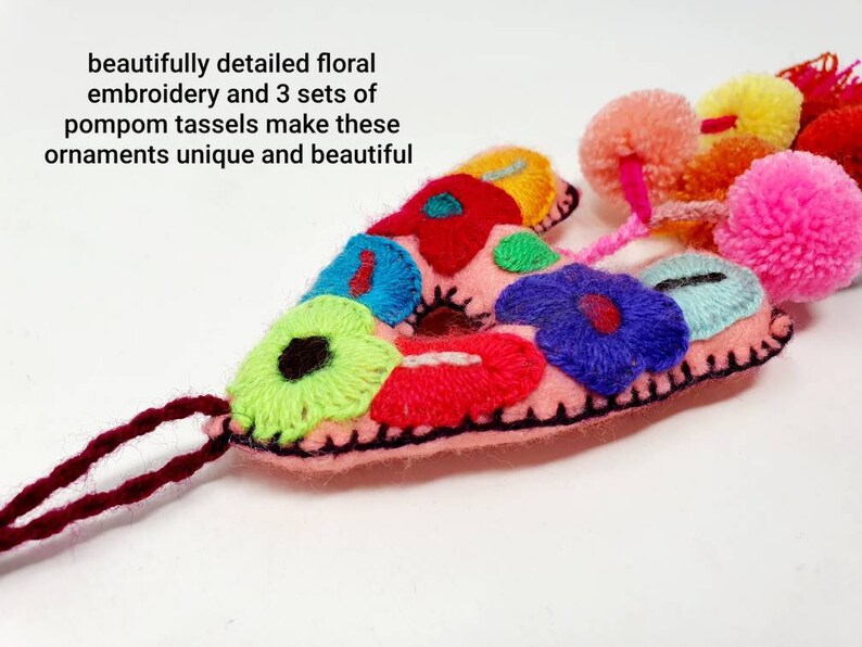 May include: Pink felt letter A with colourful floral embroidery and three sets of pompom tassels. The letter is decorated with bright pink, red, blue, green, yellow, and white flowers.