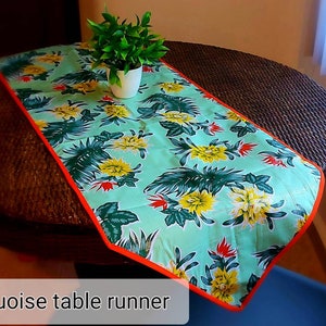 May include: Turquoise table runner with a floral pattern featuring yellow flowers and green leaves. The runner has an orange border and is on a brown wicker table.
