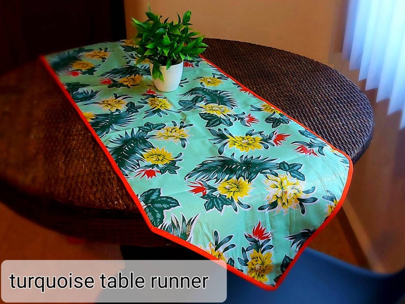 May include: Turquoise table runner with a floral pattern featuring yellow flowers and green leaves. The runner has an orange border and is on a brown wicker table.
