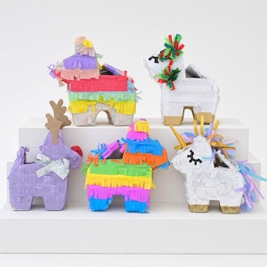 Könnte beinhalten: Fünf kleine Piñatas mit verschiedenen Designs, darunter ein lila Rentier mit silberner Schleife, ein weißes Einhorn mit goldenem Horn und drei bunte Tierfiguren, die mit Schichten aus leuchtend buntem Papierfransen verziert sind.