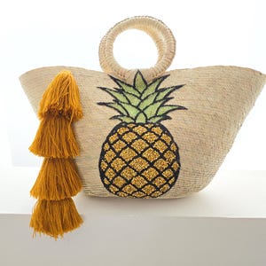 Può includere: Borsa tote in paglia intrecciata con un grande design di ananas ricamato. L'ananas presenta una corona verde e un corpo dorato strutturato. Un nappa color senape abbinata pende dal lato. La borsa ha un manico circolare.