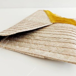 Op de afbeelding: Een geweven strooien clutch met een driehoekige klepsluiting en een felgeel kwastje. Het natuurlijk beige materiaal is geconstrueerd met een consistent visgraatpatroon over de gehele buitenkant, met een gevouwen envelopontwerp.