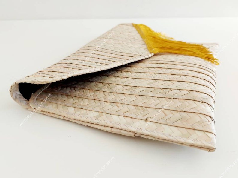 Op de afbeelding: Een geweven strooien clutch met een driehoekige klepsluiting en een felgeel kwastje. Het natuurlijk beige materiaal is geconstrueerd met een consistent visgraatpatroon over de gehele buitenkant, met een gevouwen envelopontwerp.