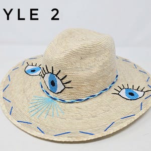 May include: A straw hat with a wide brim and a blue and white embroidered eye design. The hat is labeled "STYLE 2".