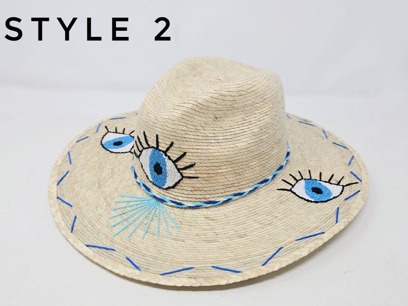 May include: A straw hat with a wide brim and a blue and white embroidered eye design. The hat is labeled "STYLE 2".