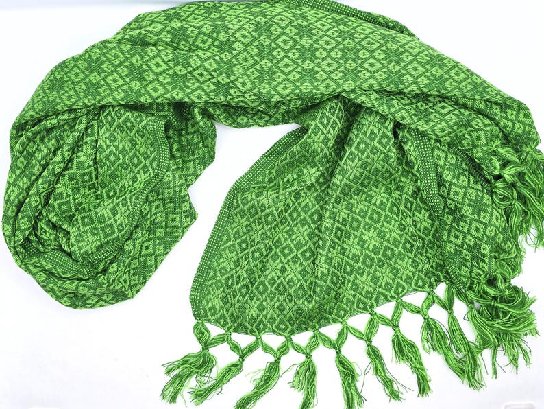 May include: A vibrant green scarf featuring a repeating geometric diamond and star pattern woven throughout the fabric. The edges of the scarf are finished with a decorative border and a series of knotted green fringe tassels along the ends.