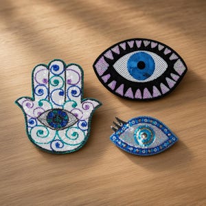Könnte beinhalten: Drei bestickte Aufnäher bestehend aus einer weißen Hamsa-Hand mit blauen und violetten Wirbeln, einem großen ovalen Auge mit blauer Iris und schwarzen Wimpern sowie einem kleineren mandelförmigen Augenaufnäher mit blauen und silbernen Pailletten.