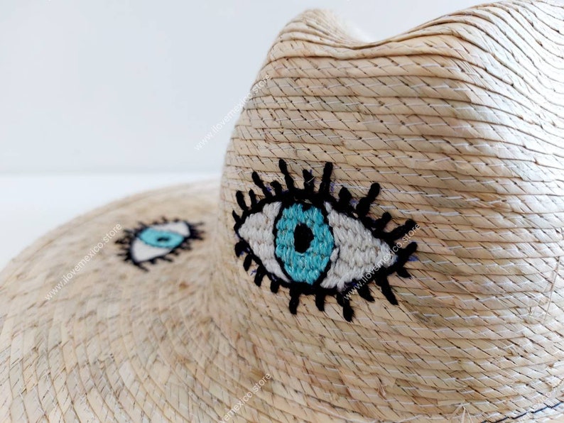 May include: A straw hat with a wide brim and two embroidered blue eyes. The eyes have white pupils and black eyelashes.