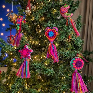 Pode incluir: Uma coleção de ornamentos de feltro coloridos pendurados numa árvore de Natal, incluindo um coração, uma estrela, uma lua crescente, uma cruz e uma forma circular, cada um com bordados florais vibrantes e longas borlas de fio.