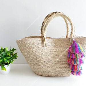 Puede incluir: Un bolso tote grande de paja con un diseño tejido y dos asas. El bolso tiene un borla colorido colgando de una de las asas.