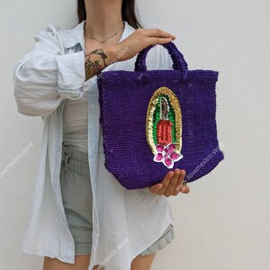 Könnte beinhalten: Lilafarbene Tragetasche mit einem bunten gestickten Bild der Jungfrau Maria. Die Tasche ist aus gewebtem Stoff und hat einen einzigen Henkel.