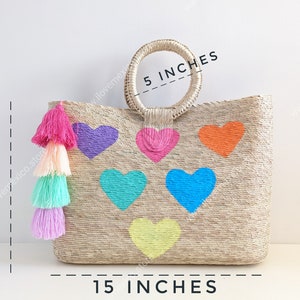 May include: A woven straw tote bag featuring six colorful heart shapes in purple, pink, orange, turquoise, blue, and yellow. A multi-colored tassel hangs from the side. Dimensions are 15 inches wide, 12 inches high, and a 5-inch handle diameter.