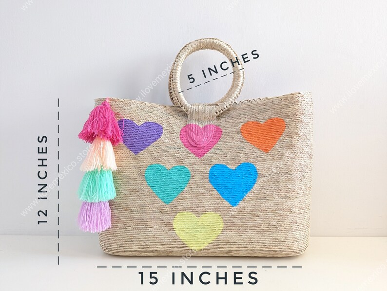 May include: A woven straw tote bag featuring six colorful heart shapes in purple, pink, orange, turquoise, blue, and yellow. A multi-colored tassel hangs from the side. Dimensions are 15 inches wide, 12 inches high, and a 5-inch handle diameter.