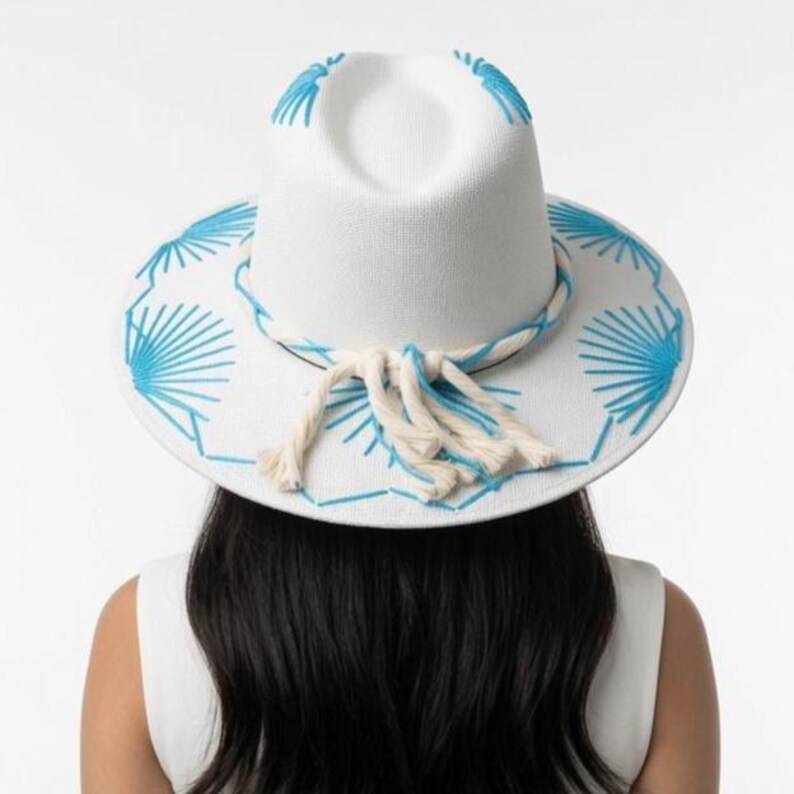 May include: A white fedora hat featuring bright blue embroidered fan-shaped patterns on the crown and wide brim. The hat is accented with a thick, braided white rope band that ties into a decorative tassel at the front center of the brim.