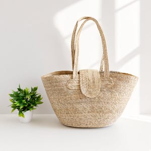May include: A light tan woven straw tote bag with two tall handles and a central flap closure. The bag features a wide, rounded base and a textured, natural fiber construction. The material is a consistent beige color throughout the entire item.
