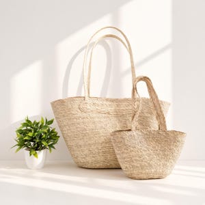 May include: Two woven straw tote bags in a light beige color, featuring a large bag with long shoulder straps and a smaller bag with shorter handles, positioned next to a small potted green plant with vibrant leaves on a flat surface.