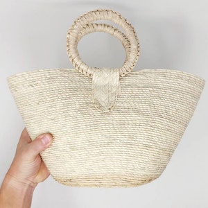 May include: A light beige woven straw handbag featuring a wide, tapered body and two circular, braided handles attached to the top center. The bag is constructed from natural fibers with a consistent horizontal coil pattern throughout the exterior.