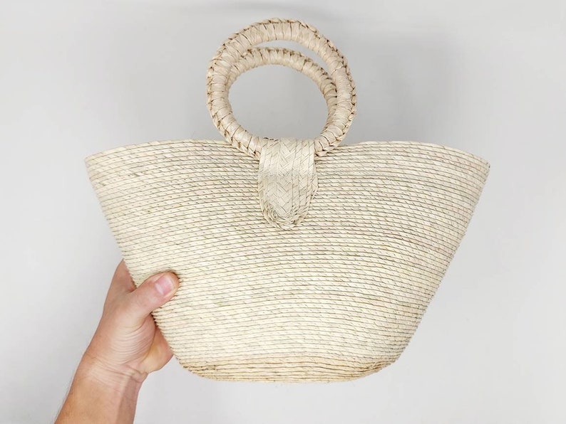 May include: A light beige woven straw handbag featuring a wide, tapered body and two circular, braided handles attached to the top center. The bag is constructed from natural fibers with a consistent horizontal coil pattern throughout the exterior.