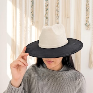 May include: A two-tone fedora hat featuring a cream-coloured crown and a wide, flat black brim. The hat has a matching cream-coloured band encircling the base of the crown, creating a distinct contrast between the light top and the dark brim.
