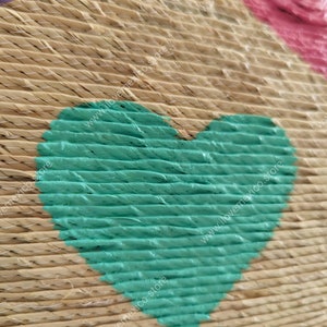 May include: A woven straw surface featuring a prominent teal heart shape painted onto the texture. The natural tan fibers are stitched together with thin white thread, and partial pink and blue heart shapes are visible at the edges.