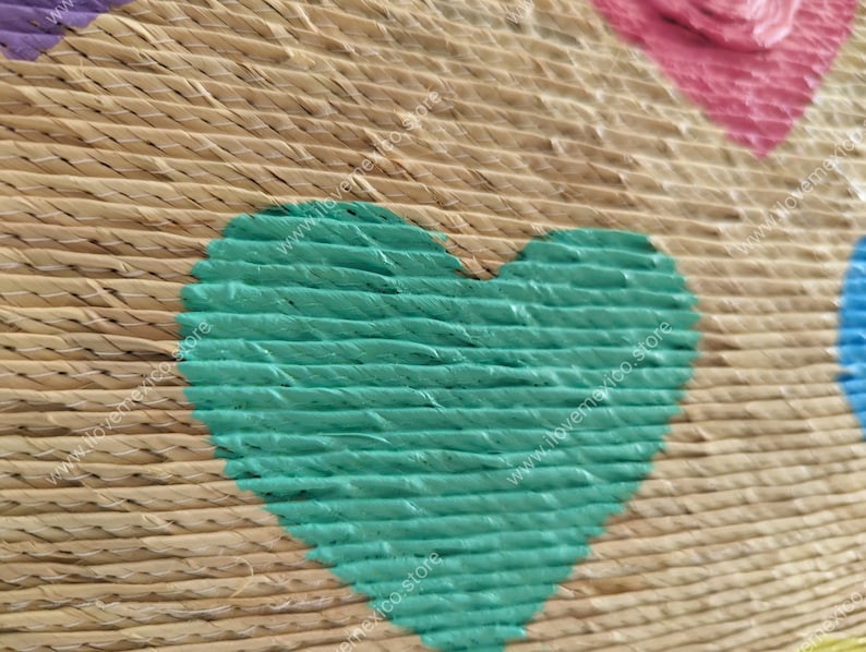 May include: A woven straw surface featuring a prominent teal heart shape painted onto the texture. The natural tan fibers are stitched together with thin white thread, and partial pink and blue heart shapes are visible at the edges.