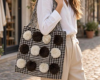 Può includere: Una borsa tote quadrata con motivo a quadri bianco e nero, decorata con pon-pon alternati marrone scuro e crema sulla parte anteriore. La borsa presenta due manici in tessuto intrecciato e una forma rettangolare strutturata con fondo piatto.