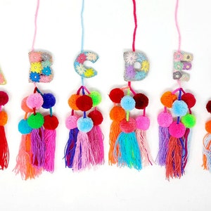 May include: Six colorful pom-pom tassels with embroidered felt letters A through F. Each tassel is made with a variety of colors, including red, orange, yellow, green, blue, and purple.