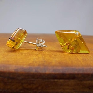 Puede incluir: Un par de pendientes de botón de ámbar. Cada pendiente presenta una piedra de ámbar geométrica y facetada con inclusiones visibles, engastada en un poste de plata. El ámbar tiene un cálido tono amarillo dorado, y los pendientes se exhiben sobre una superficie de madera.