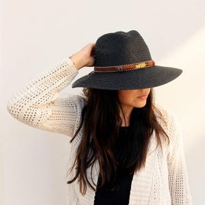 Op de afbeelding: Een donker antracietgrijze fedora-hoed met een brede rand en een geweven textuur. De hoed is voorzien van een bruine leren band versierd met kleine kraaltjes en een goudkleurige metalen veerbedel aan de voorzijde van de kroon.