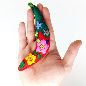 May include: A red felt chili pepper with a green felt stem and embroidered flowers in pink, yellow, blue, and green.