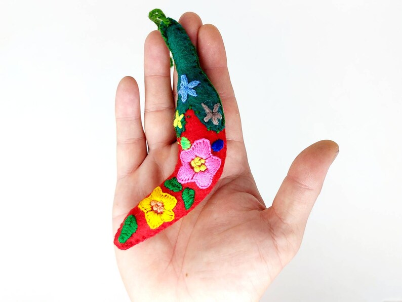 May include: A red felt chili pepper with a green felt stem and embroidered flowers in pink, yellow, blue, and green.