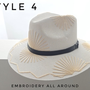 May include: A cream-colored straw fedora hat featuring beige sunburst embroidery patterns on the crown and brim, accented by a thin black band. The text STYLE 4 and EMBROIDERY ALL AROUND is visible in the image, which displays a structured design.
